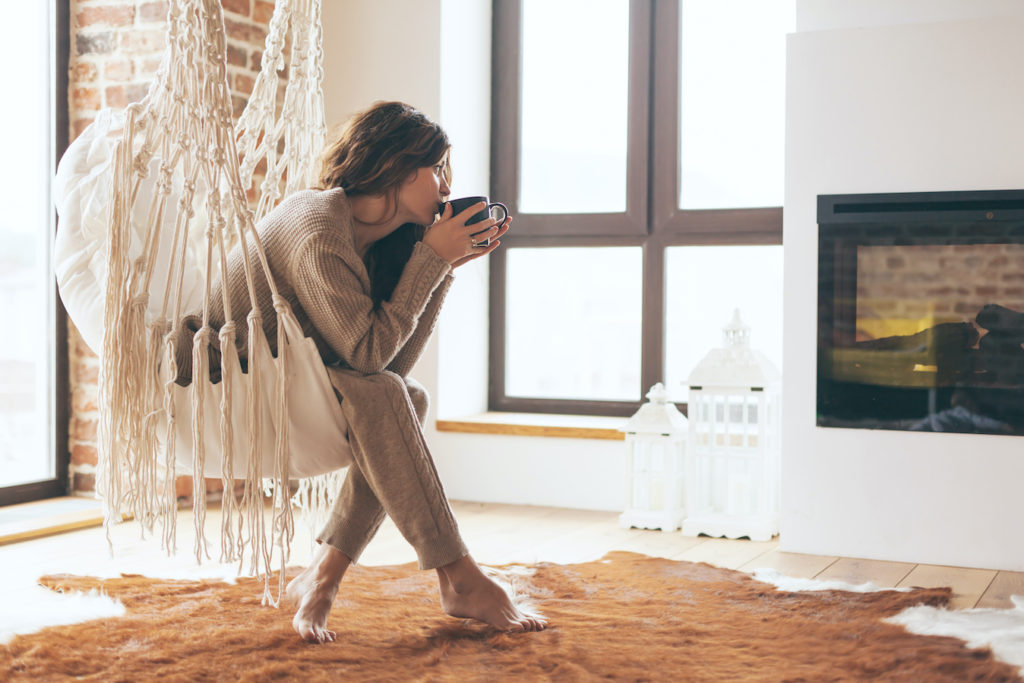 photo of a young woman sitting in a macrame hammock, sipping a warm beverage in front of a fireplace
